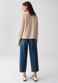 Una donna con capelli ondulati indossa un maglione beige, jeans blu a gamba larga e mocassini bianchi, in piedi contro uno sfondo grigio chiaro semplice.