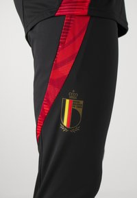 Schwarze Sporthosen mit roten Akzenten, die ein strukturiertes Design und das goldene Emblem des Königlichen Belgischen Fußballverbands aufweisen.