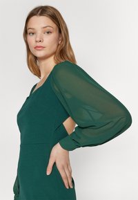 Vihreä mekko, jossa on istuva yläosa, läpikuultavat puffihihat ja avoin sivuyksityiskohta. Silkkinen kangas, jossa on pehmeä rakenne ja minimalistinen muotoilu.