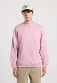 Felpa rosa chiaro con collo a crew e scritta del logo, abbinata a pantaloni beige chiaro. Presenta maniche lunghe e polsini a coste.