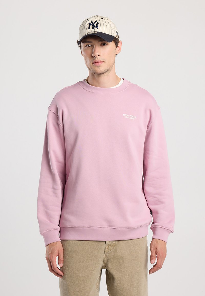 Felpa rosa chiaro con collo a crew e scritta del logo, abbinata a pantaloni beige chiaro. Presenta maniche lunghe e polsini a coste.
