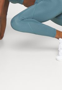Leggings ajustados en color verde azulado, hechos de un material elástico, con detalles de costura y una cinturilla alta, combinados con zapatillas deportivas blancas.