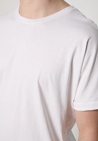 Gros plan d'une personne portant un T-shirt blanc uni à manches courtes et col rond, montrant les détails de l'épaule, du cou et du haut du bras.