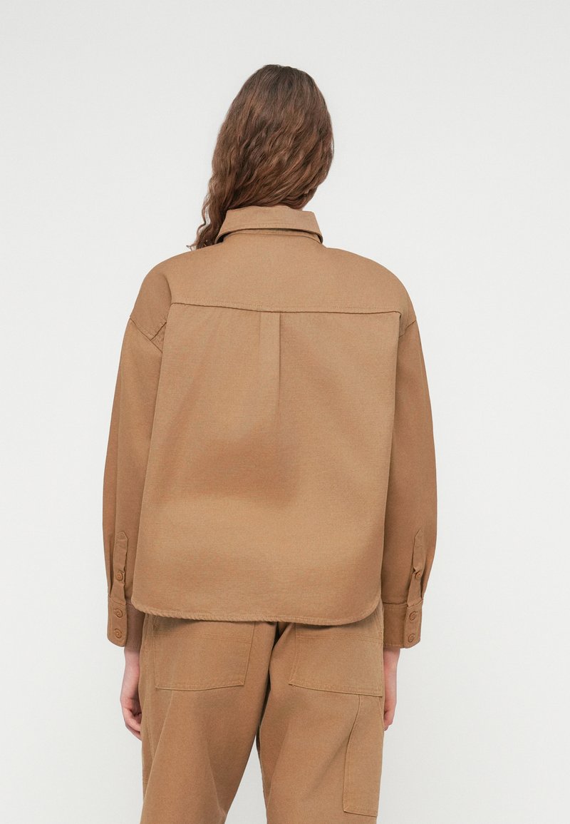 Braune Shirtjacke mit lockerer Passform, ausgestattet mit spitzem Kragen, geknöpften Manschetten und einer Rückenfalte. Hergestellt aus strukturiertem Stoff.