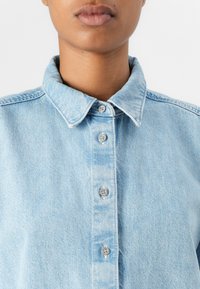 Lyseblå denimskjorte med klassisk krage, seks knapper og subtilt falmet tekstur. Glatt stoff med synlige sømdetaljer.
