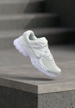 Scarpa da corsa Nike bianca con dettagli in mesh e sintetici, sospesa sopra un cornicione di cemento davanti a una scala grigia sfocata.