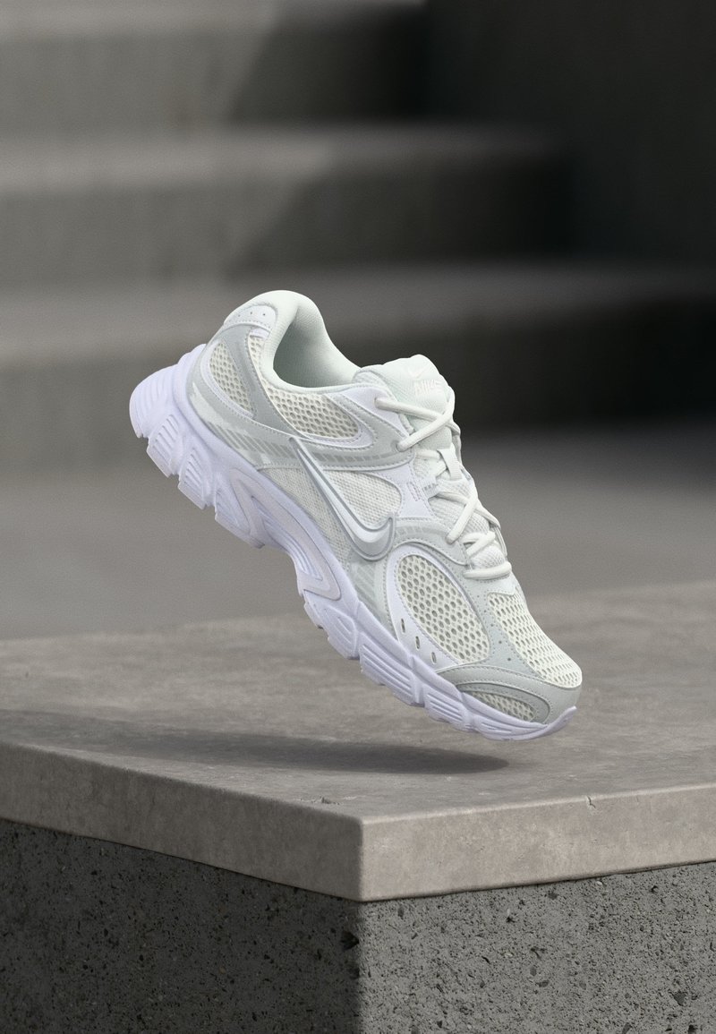 Λευκό αθλητικό παπούτσι Nike με πλέγμα και συνθετικές λεπτομέρειες, αιωρείται πάνω από μια σκυρόδετη ακμή με φόντο θολά γκρι σκαλοπάτια.