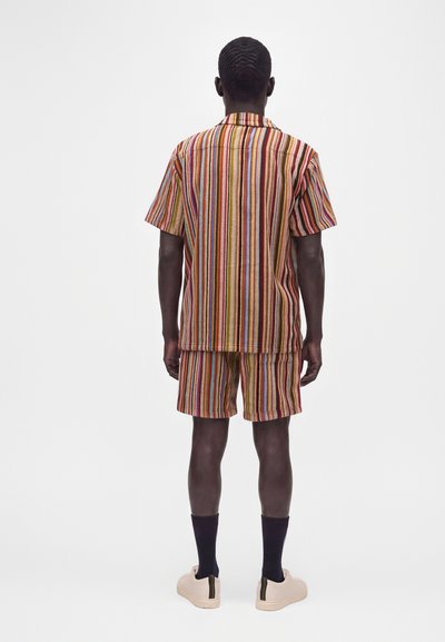 Homme debout, dos tourné, portant une chemise à manches courtes et un short assortis avec des rayures verticales multicolores et des baskets claires avec des chaussettes foncées.