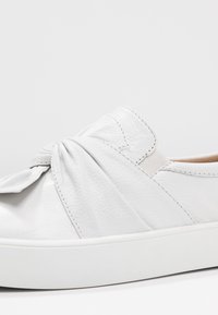 Chaussure slip-on en cuir blanc avec un design noué sur le dessus, présentant une surface texturée et une semelle en caoutchouc blanc.