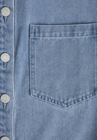 Šviesiai mėlynos džinsinės medžiagos, kuri pasižymi mygtukų užsegimu priekyje, sidabrinėmis detalėmis ir viena kišene kairėje pusėje. Horizontalus audimo raštas.