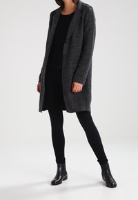 Cappotto lungo in misto lana grigio con rever a punta, due bottoni e tasche frontali. Abbinato a un top nero, leggings e stivaletti alla caviglia.