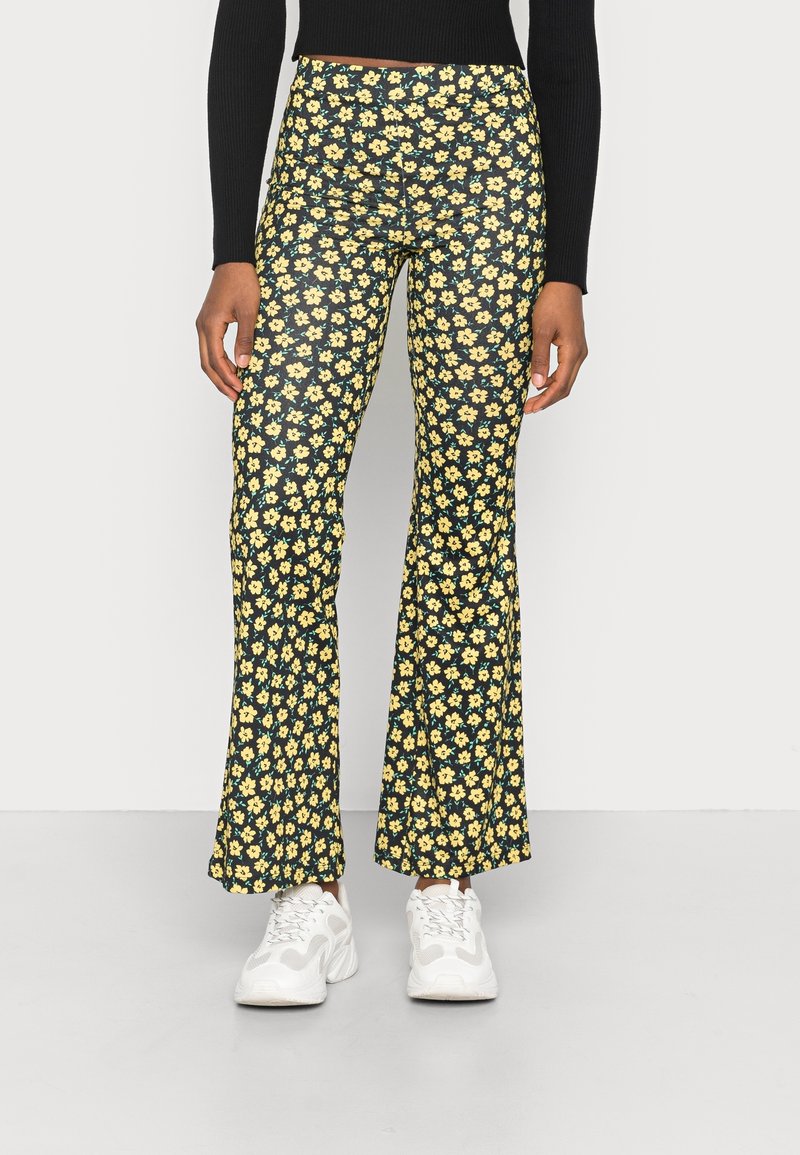Personne portant un haut noir à manches longues, un pantalon noir avec un motif floral jaune et des baskets blanches épaisses, debout sur un sol uni.