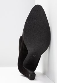 Chaussure à talon haut en daim noir avec bout pointu, vue de la semelle texturée contre un fond blanc en coin.
