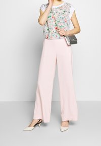 Persona vistiendo una blusa floral, pantalones anchos de un rosa claro, tacones blancos tipo slingback y llevando un pequeño bolso de cadena de plata.