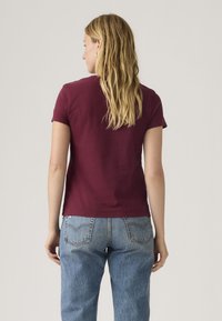 Γυναίκα που στέκεται με την πλάτη γυρισμένη, φορώντας κοντομάνικο μπορντό πουκάμισο και ανοιχτόχρωμο μπλε τζιν, μπροστά σε απλό φόντο.