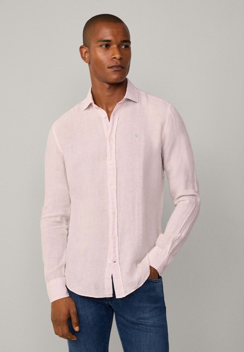 Camicia di lino rosa chiaro con maniche lunghe, chiusura frontale con bottoni e un piccolo logo blu sul petto; presenta una vestibilità rilassata e una consistenza morbida.
