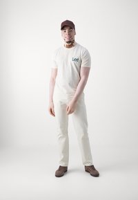 Tricou alb cu logo-ul albastru "Lee", pantaloni crema și adidași din piele de căprioară maro. Figura poartă o șapcă maro și detalii ale pielii vizibile.