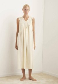 Vestido sin mangas de color crema con escote en V, que presenta detalles texturizados y un corte holgado, cayendo a media pantorrilla en una tela suave y lisa.