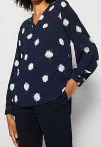 Mujer con blusa azul marino de cuello en V con manchas irregulares blancas y mangas largas, combinada con pantalones oscuros, con la mano en el bolsillo.