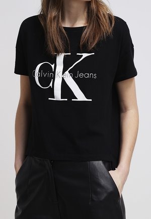 Kobieta nosząca czarną koszulkę Calvin Klein Jeans z dużym białym logo „CK” oraz czarne skórzane spodnie z rękami w kieszeniach.