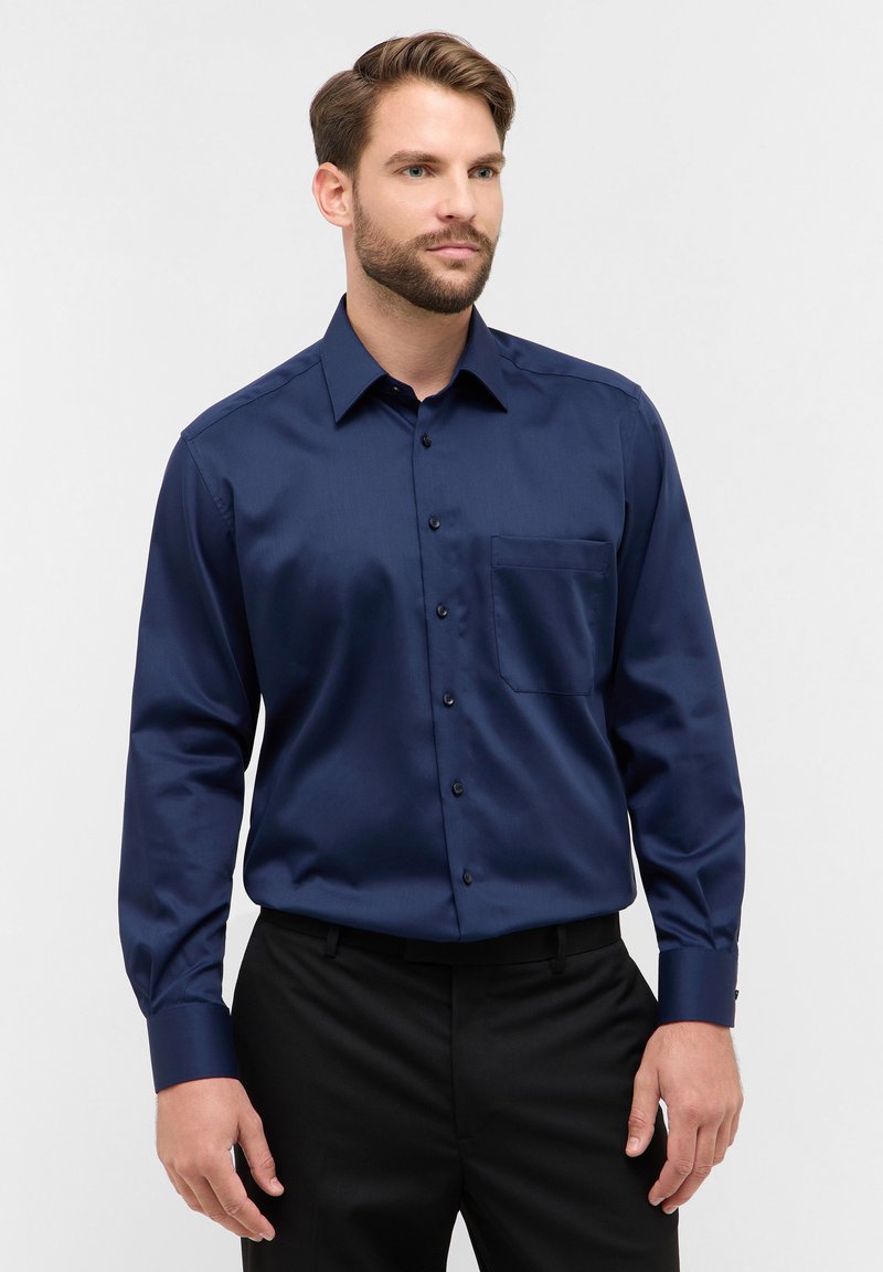 Marineblauwe longsleeve shirt van een gladde stof, met een knoopsluiting aan de voorkant, een enkele borstzak en donkere knopen. Draagt met zwarte broek.
