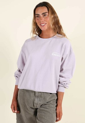 Femme souriante aux cheveux ondulés portant un sweat-shirt à manches longues lilas clair et un pantalon en jean gris sur un fond uni.