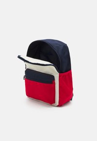 Zaino con parte superiore blu navy, corpo rosso e dettagli crema. Include una tasca frontale con zip e un compartimento principale aperto. Tessuto resistente.