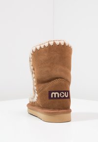 Bottine en suède marron avec des coutures crème sur le dessus, doublure moelleuse et un patch logo violet sur le côté. Semelle plate en caoutchouc.