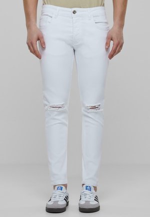 Jeans skinny albe cu rupturi distorsionate la genunchi, având închidere cu nasture, bucle pentru curea și o textură denim netedă. Purtați cu adidași gri și albi.