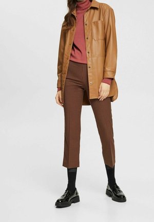 Tan faux leren overhemdjacket, roestkleurige coltrui, bruine geruite broek, zwarte sokken en glanzende zwarte schoenen. Voorzien van drukknoopsluiting en zak.