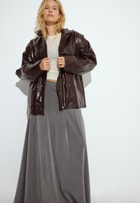 Braune Lederjacke mit Kapuze, Reißverschluss und weiten Ärmeln, kombiniert mit einem grauen, plissierten Maxikleid. Glatte Textur, minimalistisches Design.