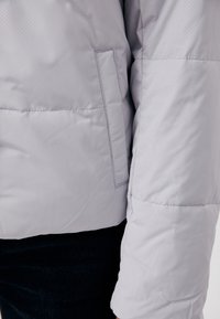 Veste matelassée gris clair avec détail de poche verticale et coutures visibles sur la manche et le côté, portée avec un pantalon foncé.