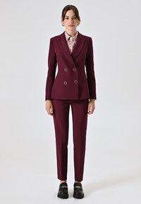 Blazer bordeaux doppiopetto con dettagli ad anello argentati, abbinato a pantaloni su misura dello stesso colore e scarpe con plateau nere. Blusa texturizzata sotto.
