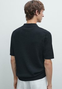 Polo shirt nero a maniche corte con lavorazione a maglia testurizzata, caratterizzato da un collo a camicia e orlo a costine, mostrato dal retro su uno sfondo neutro.