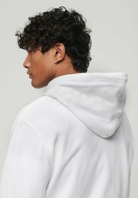 Felpa bianca con cappuccio in tessuto testurizzato, caratterizzata da un grande logo discreto sul retro e un cappuccio regolabile con cordino. Dettagli di cucitura visibili.