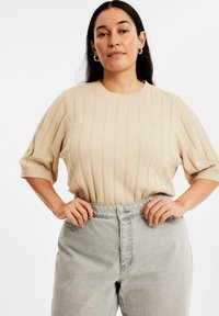 Beige ribbad stickad topp med korta ärmar, rund halsringning och subtila vertikala ränder, kombinerad med ljusgrå högmidjade jeans.