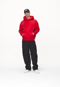 Hanorac roșu cu buzunar frontal și logo, asortat cu pantaloni cargo negri cu mai multe buzunare și adidași albi.