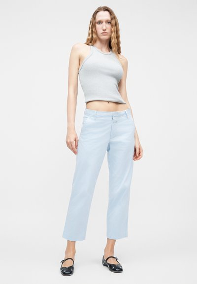 Γυναίκα με μακριά κυματιστά μαλλιά, που φοράει ανοιχτό γκρι αμάνικο crop top, ανοιχτό μπλε παντελόνι σε ίσια γραμμή και μαύρα σανδάλια με λουράκια, στέκεται πάνω σε λευκό φόντο.