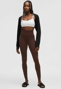 Leggings marron taille haute en tissu élastique, associés à un crop top blanc et des sandales noires à bout ouvert. Long cardigan noir.
