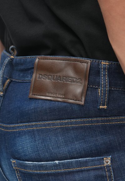 Jeans de mezclilla azul con una etiqueta de cuero marrón de DSQUARED2 en la cinturilla, que presentan costuras amarillas y un bolsillo con un remache de metal.