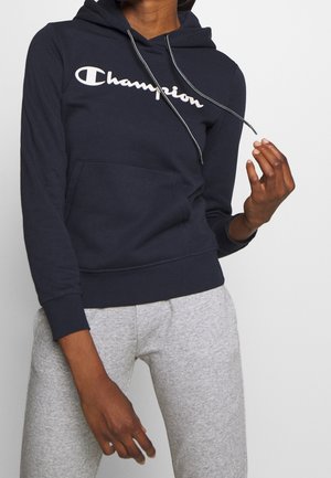 Person trägt einen marineblauen Champion-Hoodie mit weißem Logo, kombiniert mit hellgrauen Jogginghosen, und zieht mit der rechten Hand am Kordelzug des Hoodies.