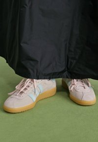 Light grey suede sneakers with blue accents, round toe, and beige rubber soles, partially obscured by a black skirt on green flooring.