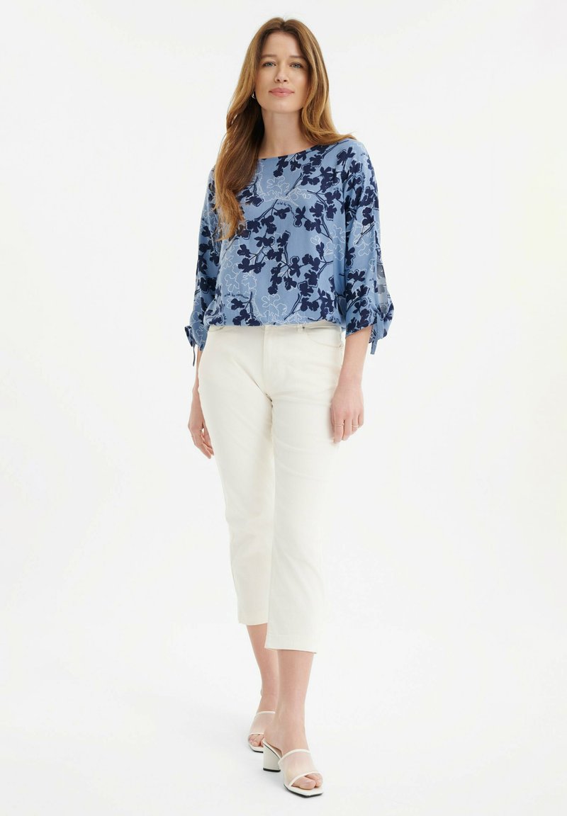 Niebieska bluzka w kwiaty z rękawami do 3/4, zdobiona granatowymi kwiatami na jasnoniebieskim tle; zestawiona z białymi, krótkimi spodniami i butami.