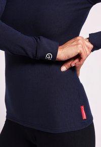 Camiseta de manga larga en color navy con un diseño ajustado, que presenta un logo en el puño y una etiqueta roja en el dobladillo, confeccionada con material elástico.