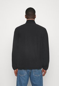 Černá zipová bunda s vysokým límcem a texturovaným povrchem. Obsahuje kontrastní barevný detail kolem límce, v kombinaci s modrými džíny.