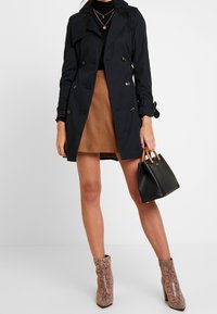 Femme portant un trench noir, un col roulant noir, une jupe camel, des bottines à imprimé serpent et tenant un sac à main noir avec des poignées en bois.