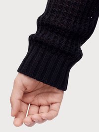 Rechte Hand mit entspannten Fingern, die einen schwarzen langärmeligen, gestrickten Pullover mit Bündchen auf hellem Hintergrund trägt.