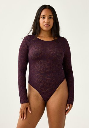 Donna con capelli lunghi e scuri che indossa un body viola in pizzo a maniche lunghe, in piedi su uno sfondo chiaro e semplice.