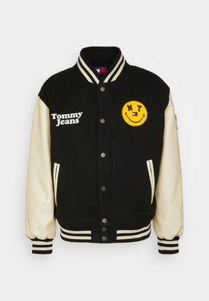 Sort og creme varsityjakke med stribede manchetter, forreste trykknapper, "Tommy Jeans" tekst og et gult smiley-patch med "NY" og et logo.