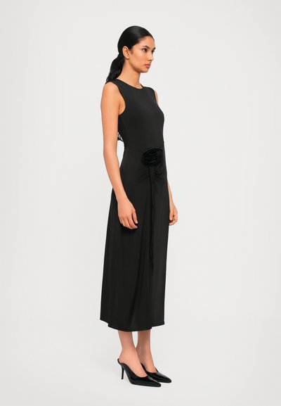 Svart ärmlös klänning med en figurnära bodice, flödande kjol och en samlad blommig detalj vid midjan. Modell som bär svarta högklackade skor.
